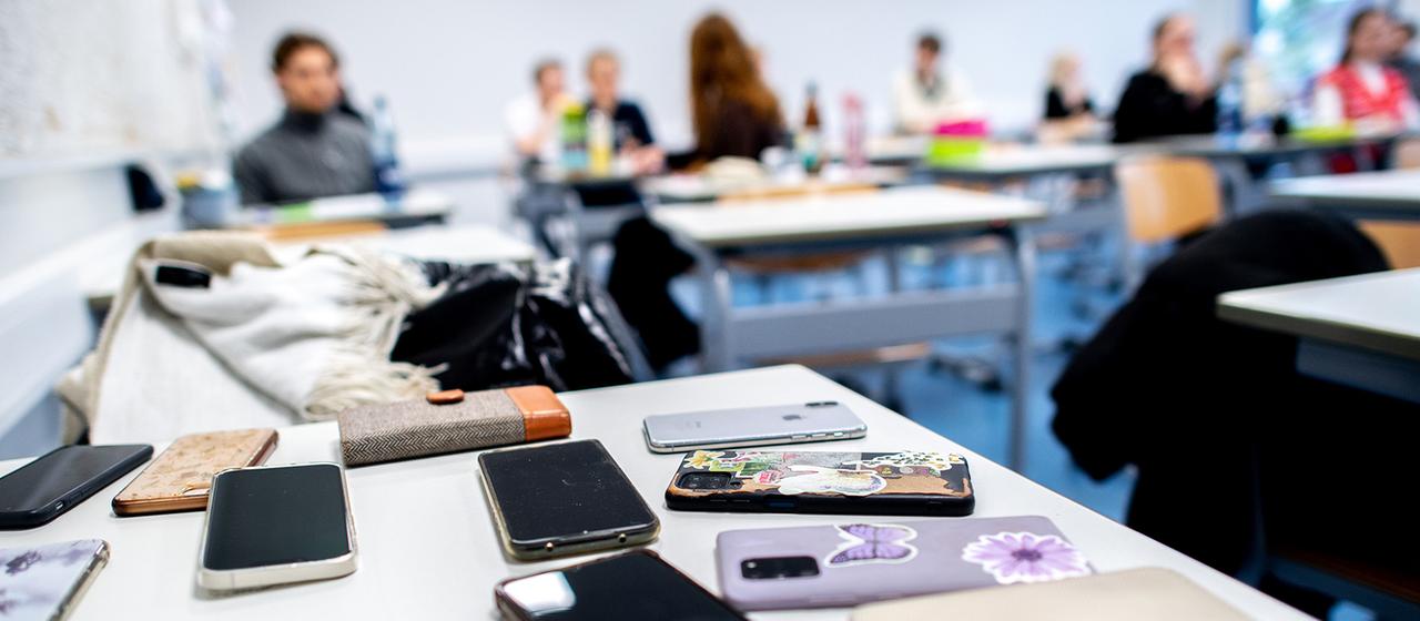 In einem Unterrichtsraum einer Schule liegen zahlreiche Smartphones auf einem Tisch.