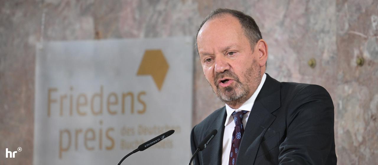 Philippe sands in der Paulskirche: Eine Person in dunklem Anzug und Krawatte steht hinter einem steinernen Rednerpult und spricht in zwei Mikrofone. Im Hintergrund ist ein Schild mit der Aufschrift "Friedenspreis des Deutschen Buchhandels" und dem zugehörigen Logo zu sehen. Die Wand dahinter besteht aus hell gemasertem Naturstein.