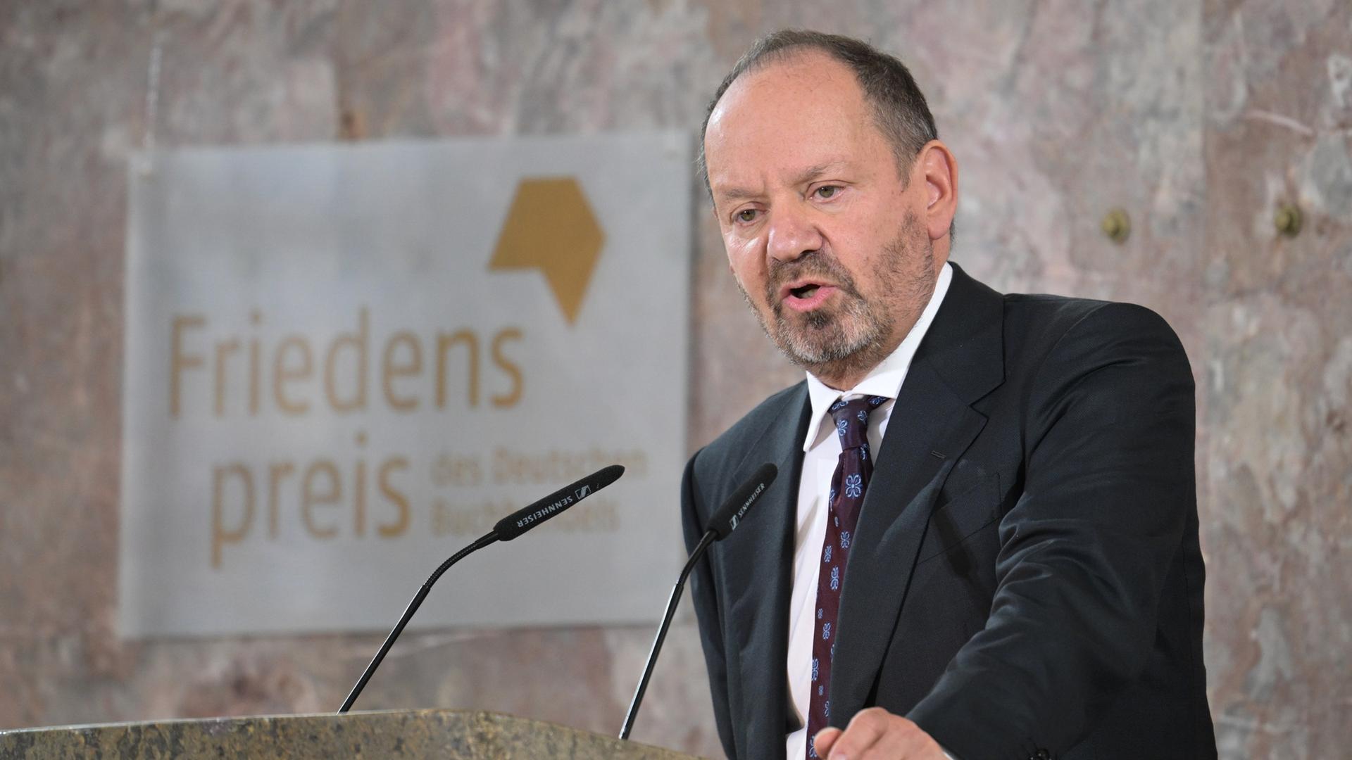 Philippe sands in der Paulskirche: Eine Person in dunklem Anzug und Krawatte steht hinter einem steinernen Rednerpult und spricht in zwei Mikrofone. Im Hintergrund ist ein Schild mit der Aufschrift 