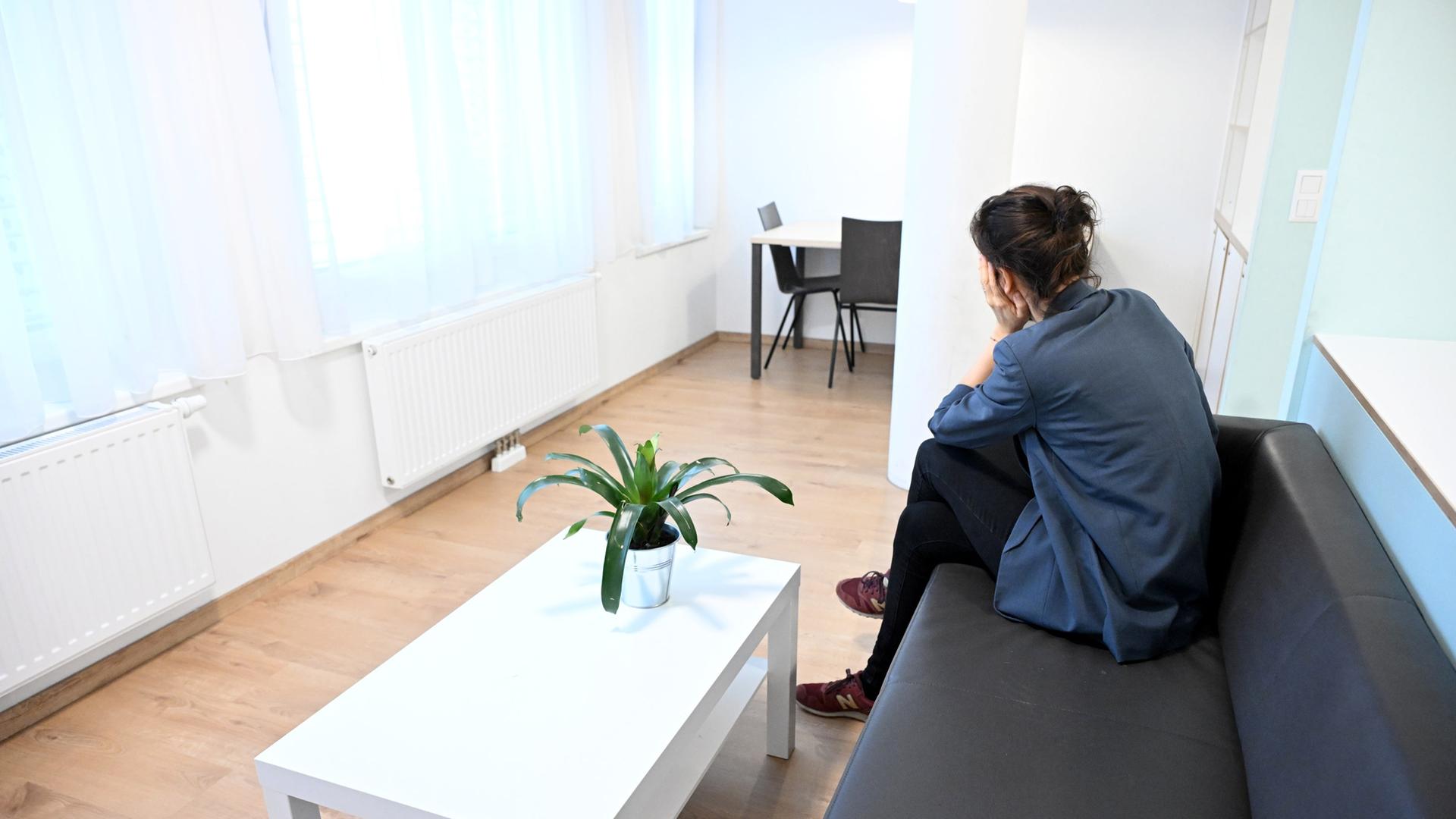 Eine dunkelhaarige Frau sitzt in einem spärlich möblierten Raum mit weißen Wänden auf einem Sofa und wendet sich von der Kamera ab, so dass sie nur von hinten zu sehen ist | picture-alliance/dpa