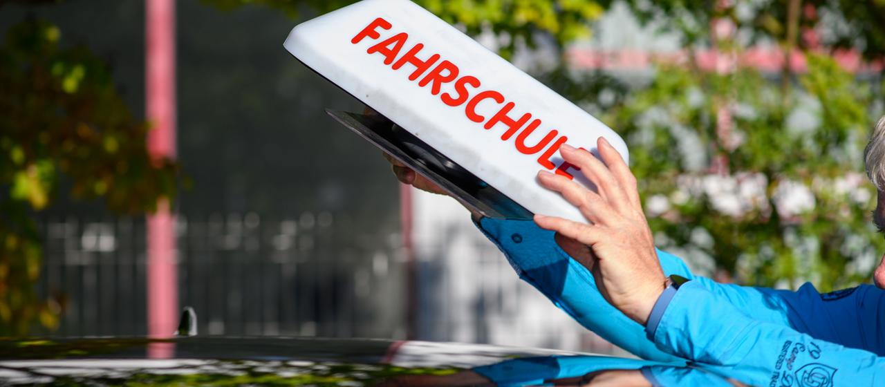"Fahrschule" ist auf einem Schild zu lesen das ein Fahrlehrer auf das Dach seines Autos stellt.
