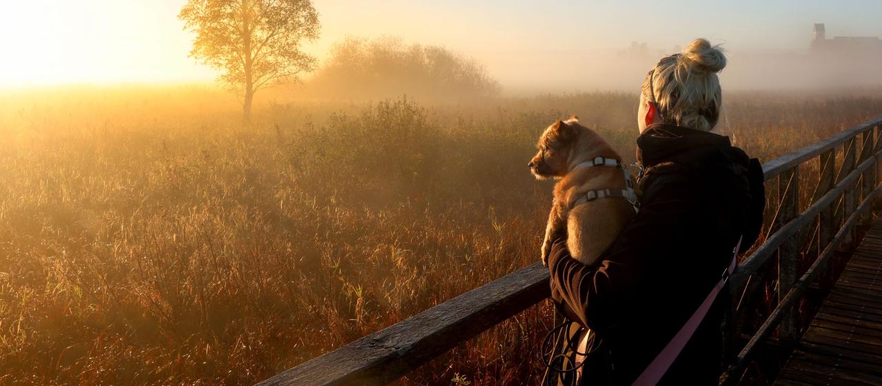 Eine Frau mit ihrem Hund bei Sonnenaufgang am Federsee, 	Baden-Württemberg.