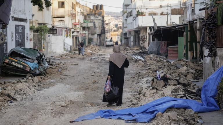 Eine Frau mit Kopftuch läuft durch eine Straße in Dschenin. Den Straßenrand säumen Schutt und Autowracks.