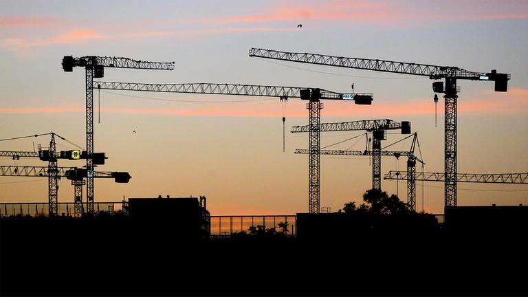 Baukräne zum Bau von Mehrfamilienhäusern zeichnen sich vor dem von Sonnenaufgang rötlich verfärbten Morgenhimmel ab. 