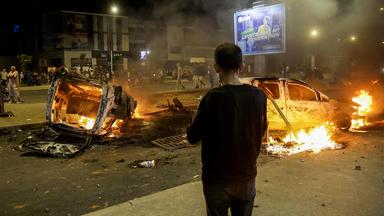 Ein Mann steht vor brennenden Autos auf einer Straße.