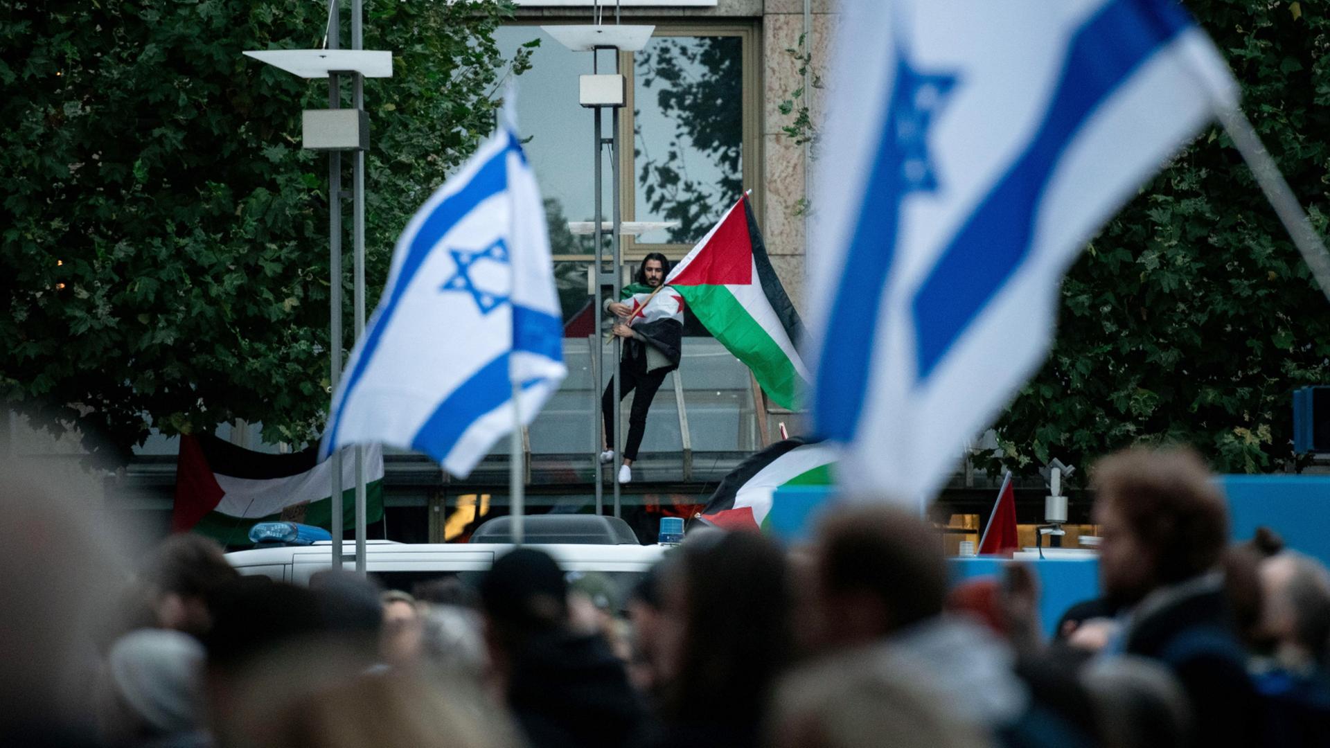 Ein Teilnehmer einer pro-palästinensischen Demonstration ist mit einer Palästina-Fahne hinter einer pro-israelischen Demonstration zu sehen. (Archivbild vom 18.10.2025) | dpa