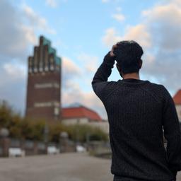 Mensch, der sich an seinen Kopf greift - von hinten fotografiert. Im Hintergrund unscharf ein urbanes Umfeld mit der "Mathildenhöhe".