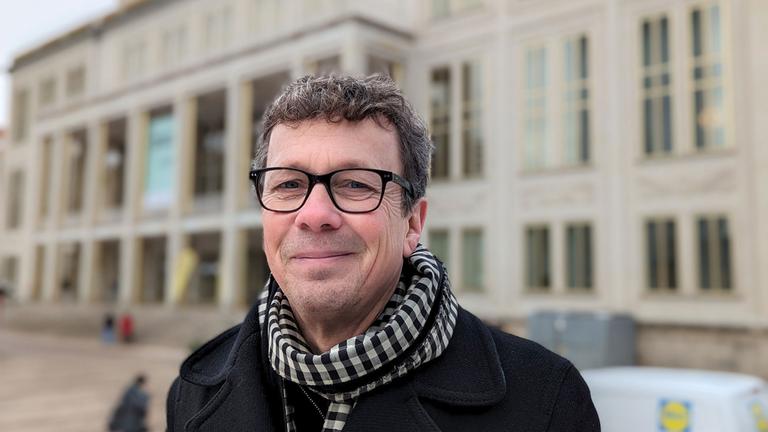 Ein Mann um die fünfzig mit schwarzer Wolljacke, schwarz-weißem Schal und schwarz umrandeter Brille lächelt in die Kamera. Er hat kurzes braunes Haar und steht vor einem Opernhaus.