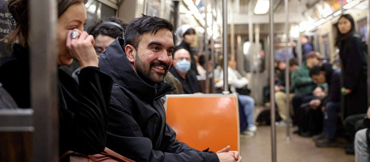 Zohran Mamdani sitzt lächelnd in der U-Bahn.