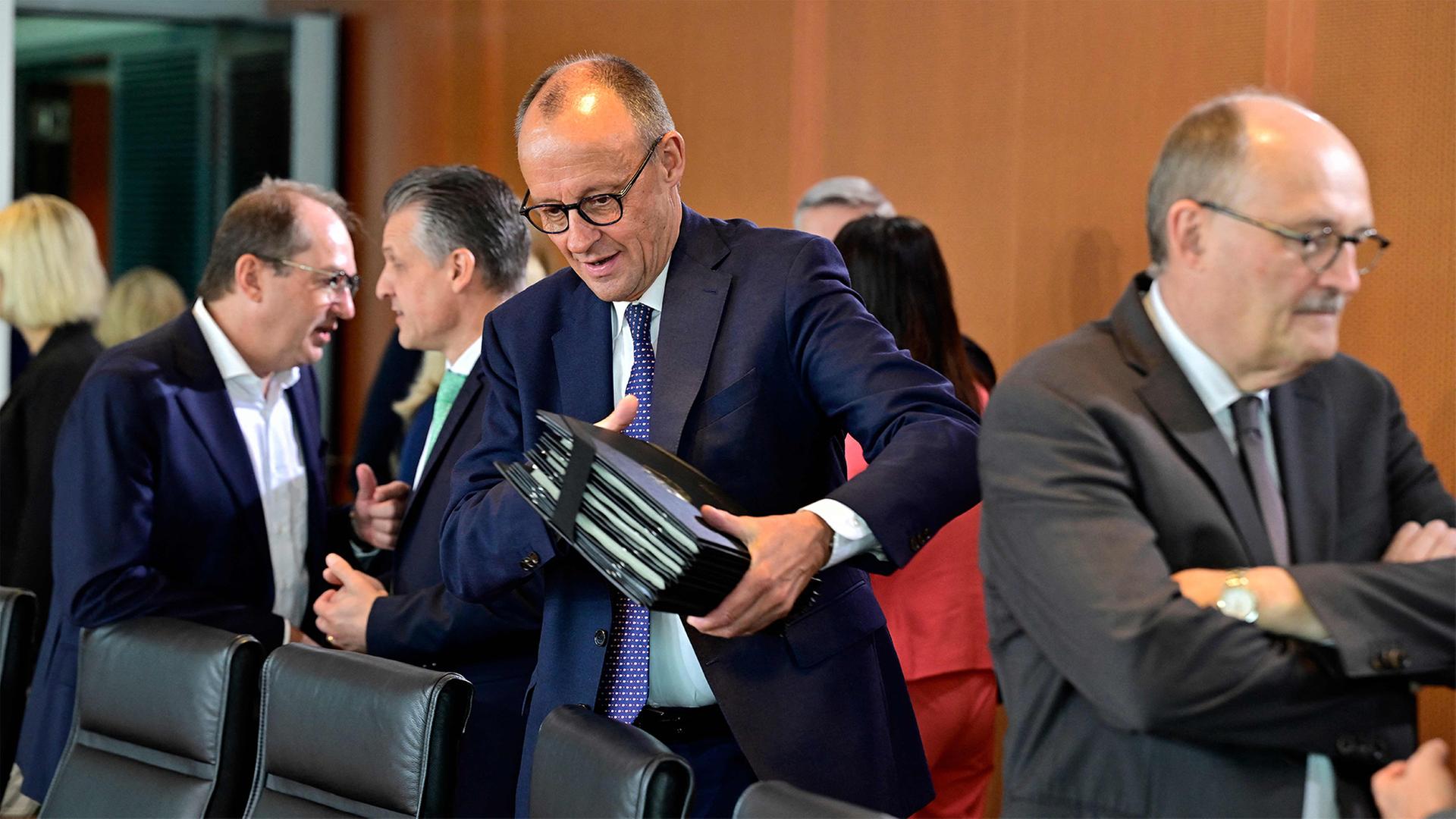 Friedrich Merz Im  Konferenzraum des Bundeskabinetts. Er  hält mehrere schwarze Aktenmappen in den Armen. Weitere Personen stehen im Hintergrund und unterhalten sich. | AFP