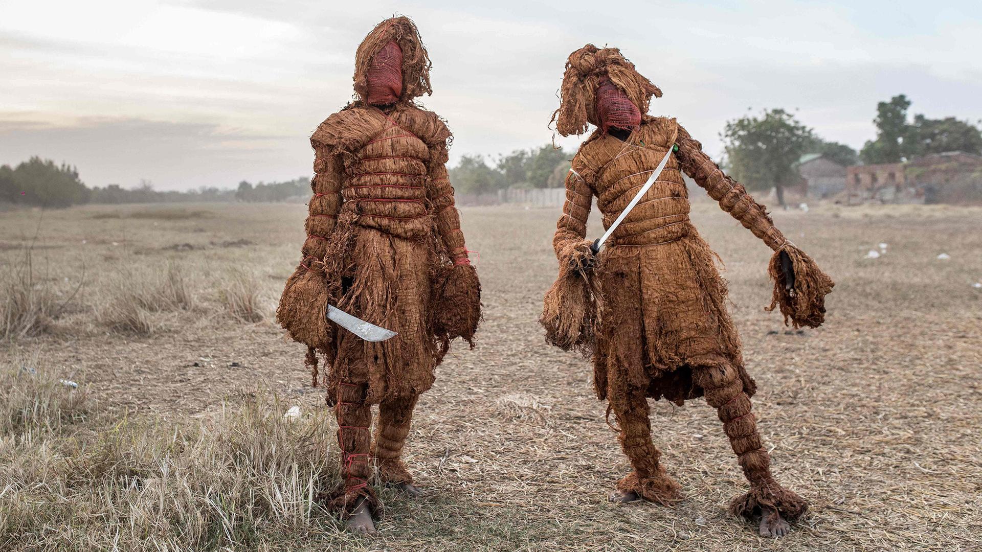 Zwei Personen stehen auf einem Feld und tragen traditionelle Verkleidungen aus Pflanzenfasern und roten Stoffmasken. Beide halten eine Machete in der Hand. | AFP