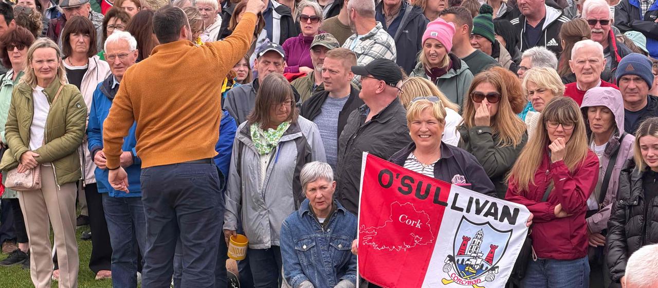 Männer und Frauen unterschiedlichen Alters stehen in einer Menge zusammen, eine Frau hält eine Fahne mit dem Namen O'Sullivan hoch.