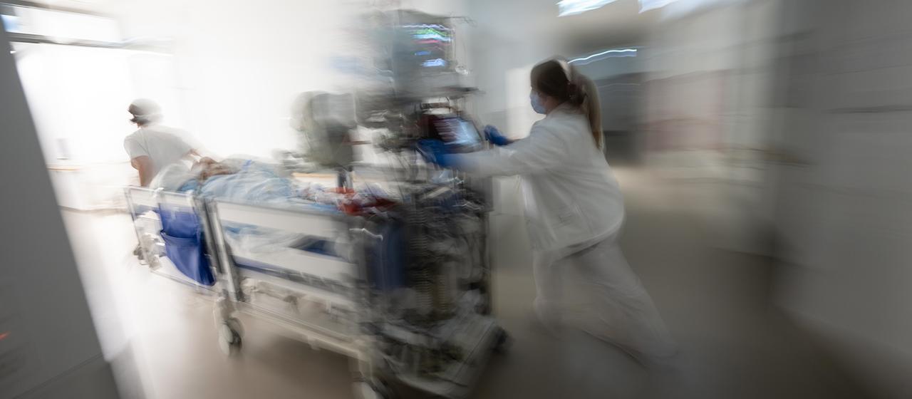Eine Krankenschwester fährt einen Intensivpatienten in einem Krankenhausbett durch einen Krankenhausflur. | dpa Eine Krankenschwester fährt einen Intensivpatienten in einem Krankenhausbett durch einen Krankenhausflur.