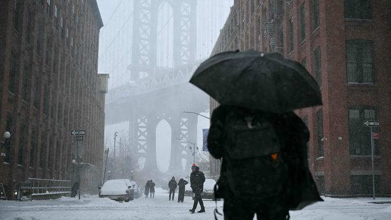 Menschen gehen durch eine verschneite Straße in New York, im Hintergrund ist die Manhattan Bridge zwischen zwei Backsteingebäuden zu sehen.