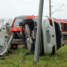 Ein Auto liegt auf der Seite neben einer zerstörten Schranke an einem Bahnübergang. Im Hintergrund steht ein Zug auf den Gleisen. 