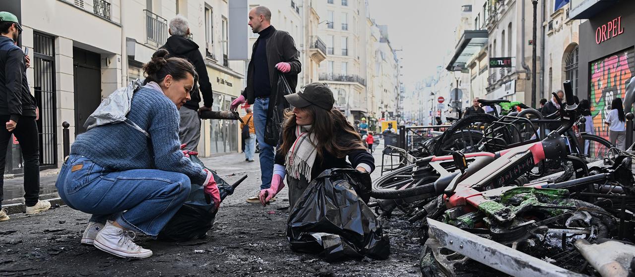 Menschen räumen auf einer Straße neben ausgebrannten Fahrrädern Trümmer weg.