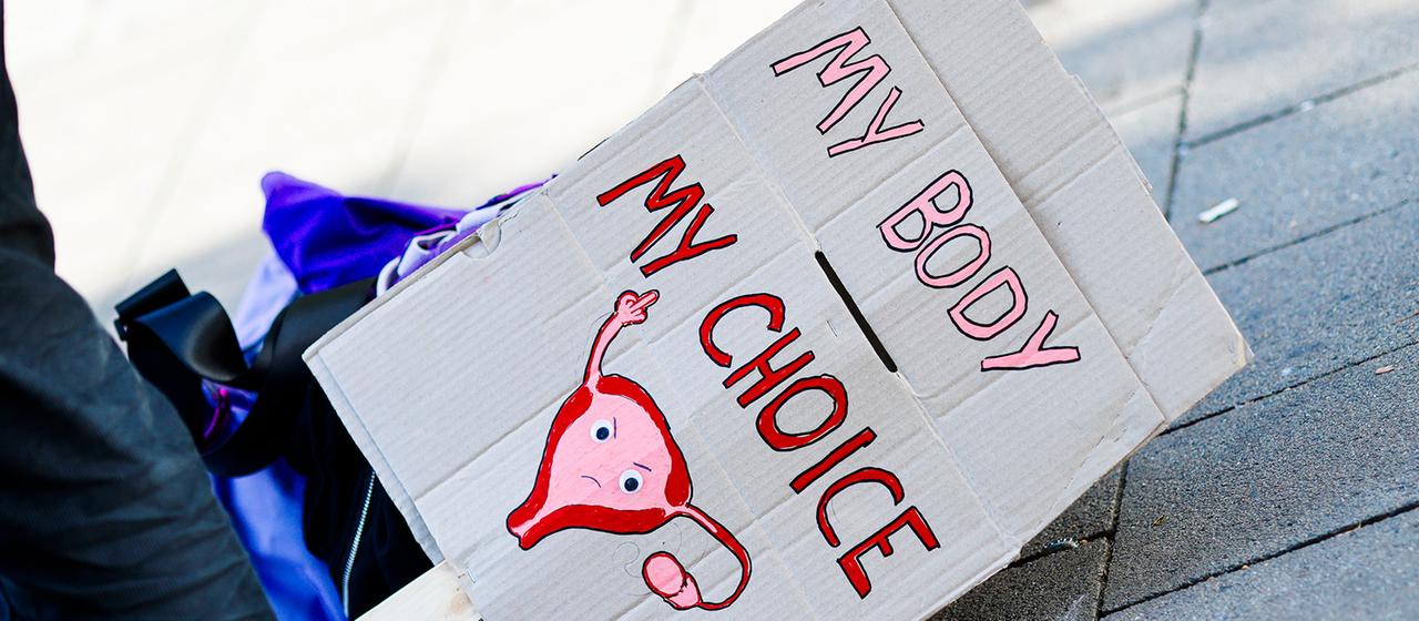 Ein Pappschild mit der Aufschrift "MY BODY MY CHOICE" und einer Zeichnung einer Gebärmutter mit erhobenem Arm und ausgestrecktem Finger.