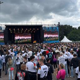 In Oberhausen können bis zu 40.000 Menschen das erste Spiel der deutschen Nationalmannschaft bei der WM 2026 sehen. Auf großen Leinwänden ist zu sehen, wie die deutschen Spieler die Nationalhymne singen. Der Platz vor den Leinwänden ist mit Fans gefüllt.