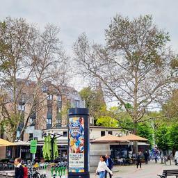 Der Frankfurter Merianplatz mit einer Litfasssäule und einem Café im Zentrum, im Vordergrund Menschen, im Hintergrund Häuser und zwei Platanen mit teils grünen, teils abgestorbenen Blättern