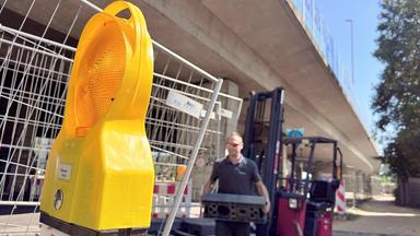 Unter einer massiven Betonbrücke steht ein gelbes Warnlicht, rechts dahinter ein Bauarbeiter in dunkler Arbeitskleidung, der ein Gewicht trägt.