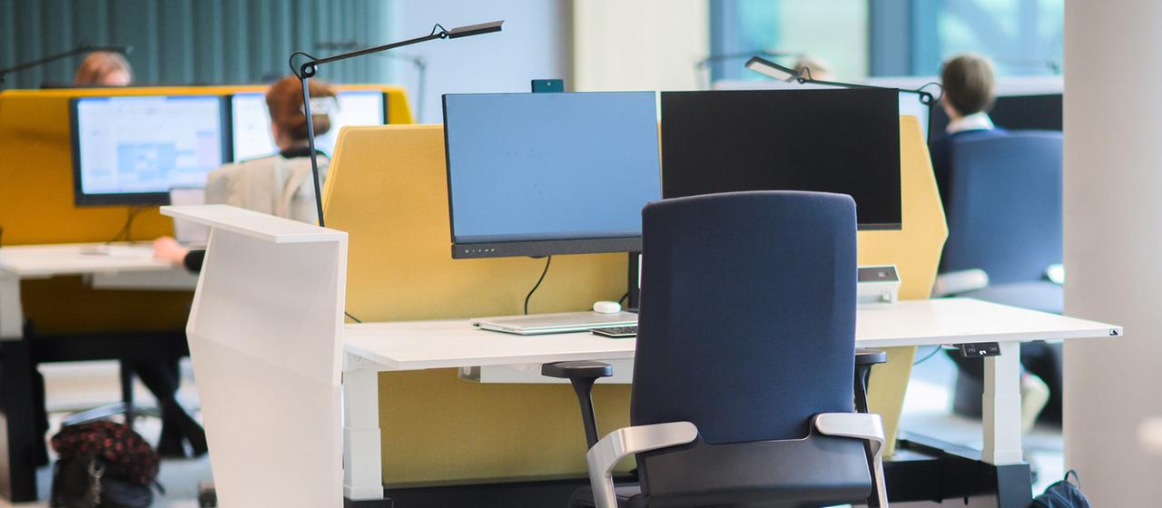 Mehrere moderne Arbeitsplätze in einem Großraumbüro mit Computermonitoren, Schreibtischen und Bürostühlen, einige Personen sitzen an den hinteren Arbeitsplätzen.