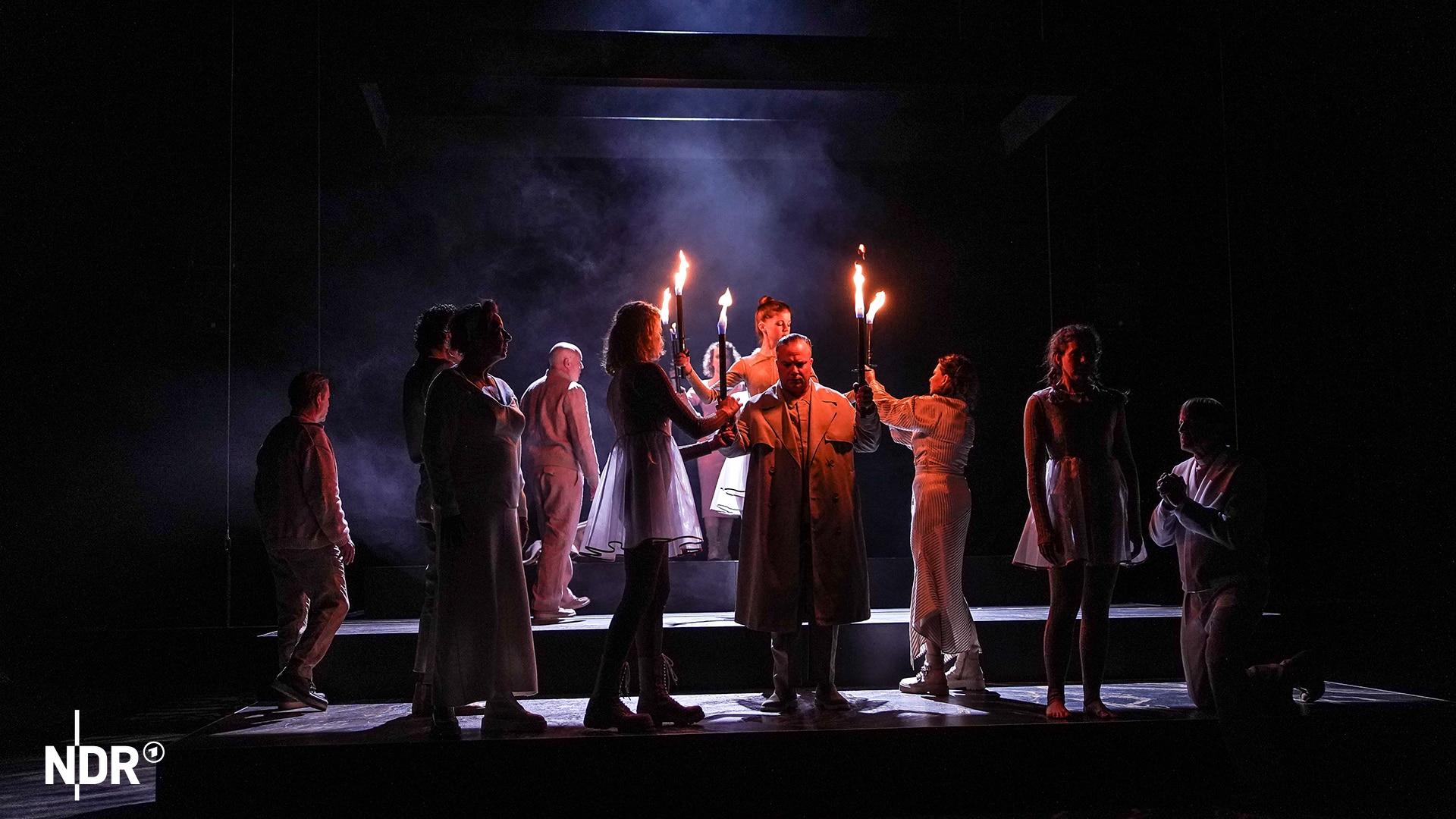 Eine Gruppe Menschen, beleuchtet vom roten Schein brennender Fackeln, steht auf einer Theaterbühne. | Schauspielhaus Kiel Eine Gruppe Menschen, beleuchtet vom roten Schein brennender Fackeln, steht auf einer Theaterbühne.