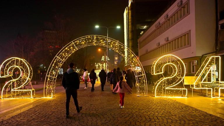 Menschen gehen in Pristina durch ein beleuchtetes Tor, das die Jahreszahl 2024 anzeigt.