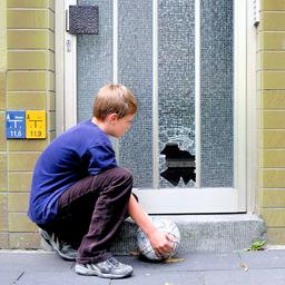 Ein Junge mit einem Fußball betrachtet die eingeschossene Glasscheibe an einer Haustür.