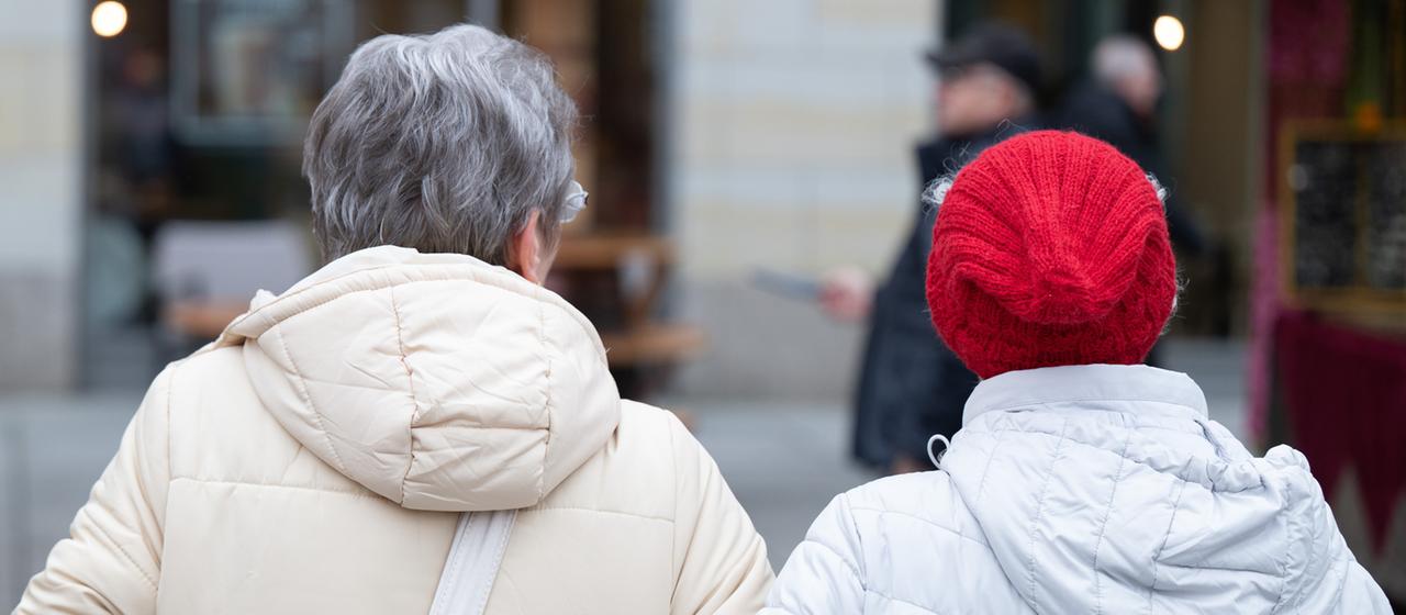 Zwei ältere Frauen gehen eingehakt durch eine Einkaufsstraße.