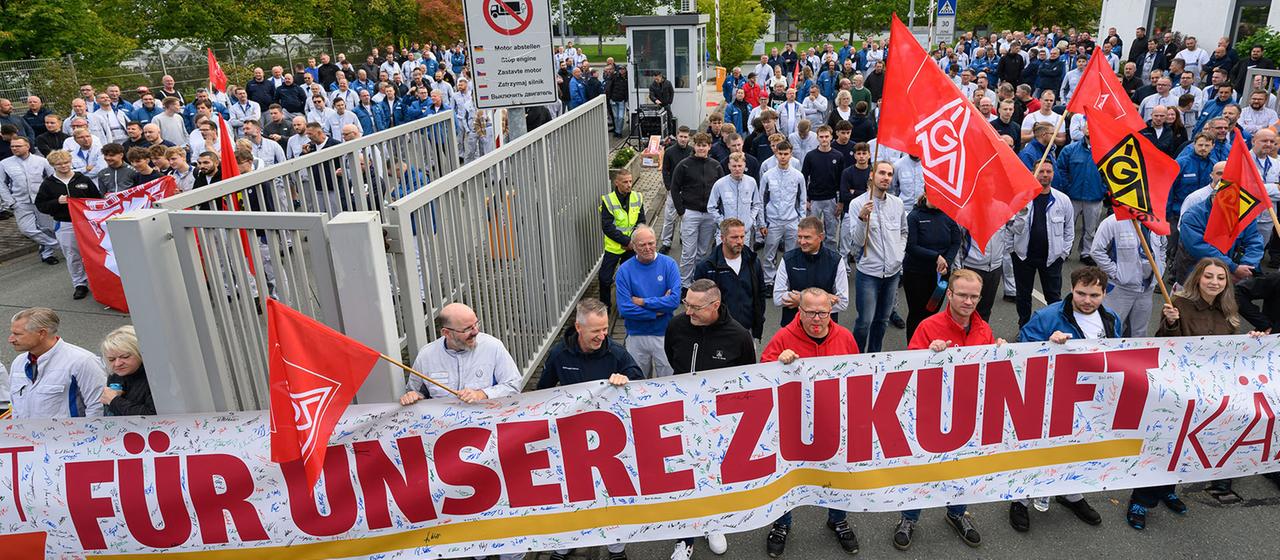 Menschen mit einem großen Banner und IG-Metall-Flaggen demonstrieren vor einem Werkstor.