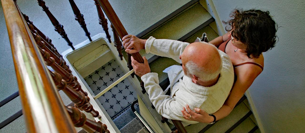 Eine Frau unterstützt einen älteren Mann beim Treppensteigen in einem Treppenhaus.