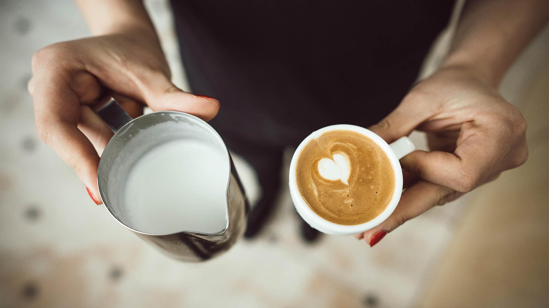Eine Barista hält eine Tasse Kaffee und ein Kännchen Milch in den Händen. | picture alliance / Zoonar