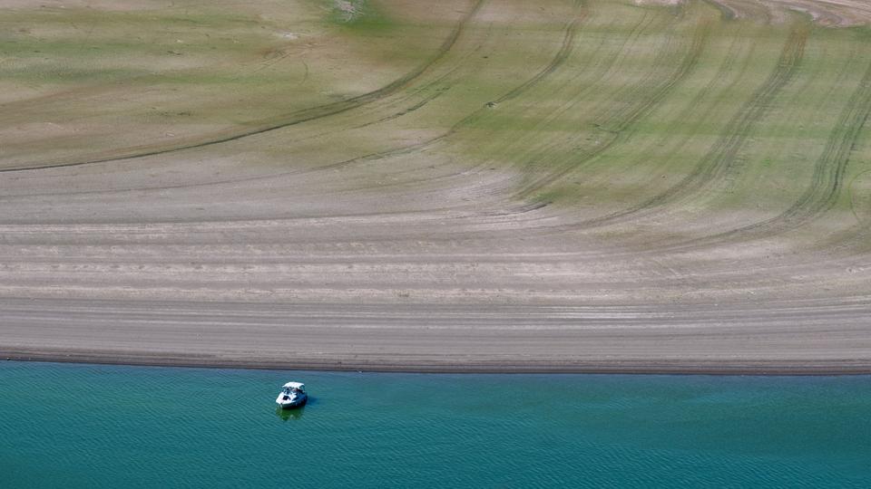 Ein Boot fährt auf dem Edersee im hessischen Waldeck unterhalb einer großflächig trockengefallenen Uferzone. 