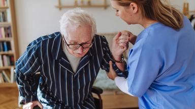 Eine Pflegekraft hilft einem alten Mann aus dem Rollstuhl aufzustehen.