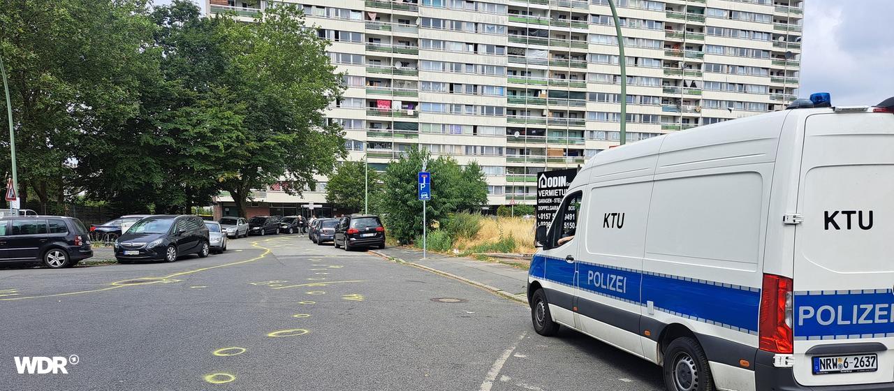 Ein Fahrzeug der Kriminaltechnischen Untersuchung steht am Straßenrand. Im Hintergrund ist der Weiße Riese in Duisburg, ein weißes Hochhaus zu sehen. Auf der Straße sind gelbe Kreide-Markierungen zu erkennen.