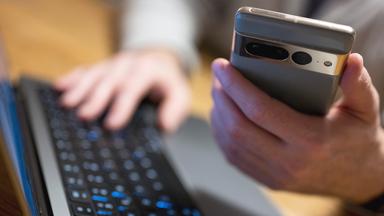 Ein Mann sitzt an einem Laptop und hält ein Mobiltelefon in der Hand.