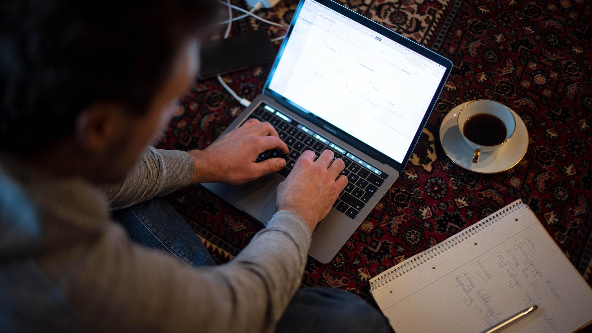 Ein Mann sitzt im Homeofficean seinem Laptop auf einem Teppich. | picture alliance/dpa