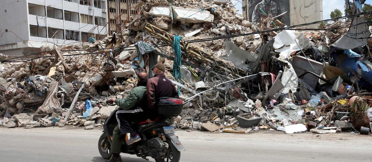 Menschen fahren auf einem Motorroller an zerstörten Gebäuden im Süden Libanons vorbei.