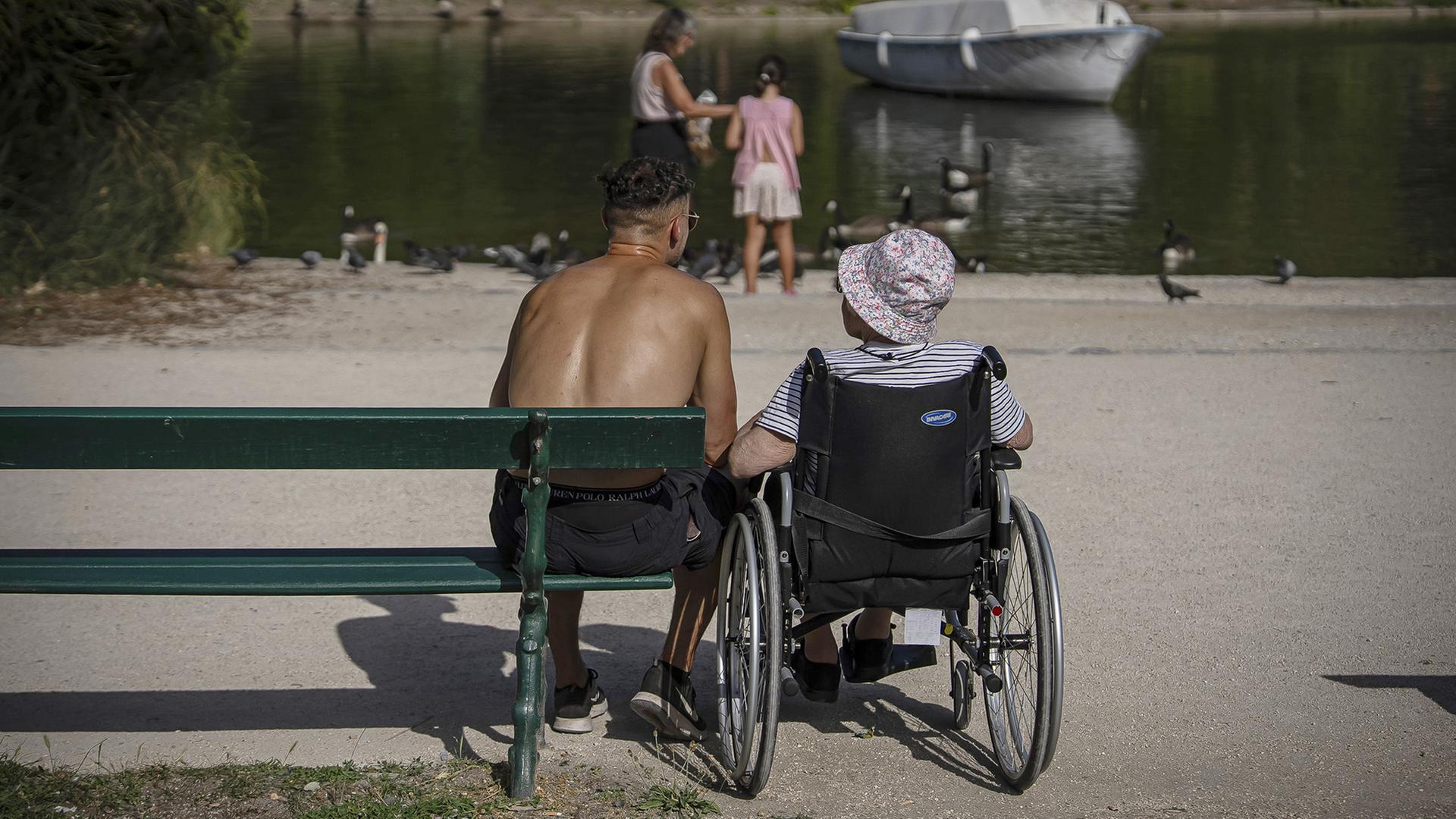 Zwei Personen sitzen vor einem Teich, eine ist im Rollstuhl, die andere Person sitzt auf einer Bank. | AP
