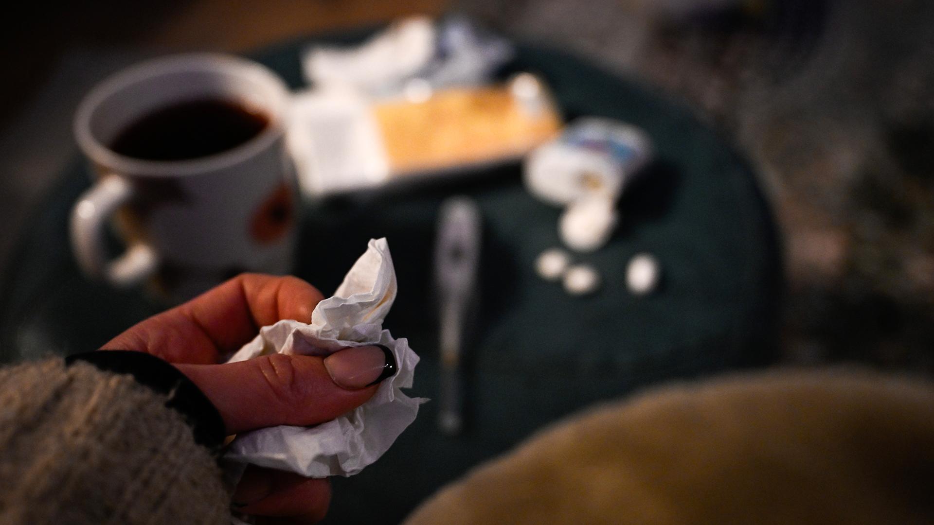 Eine Hand mit einem Taschentuch ist vor einem Tisch mit Tee, Fieberthermometer und Medikamenten zu sehen. | dpa