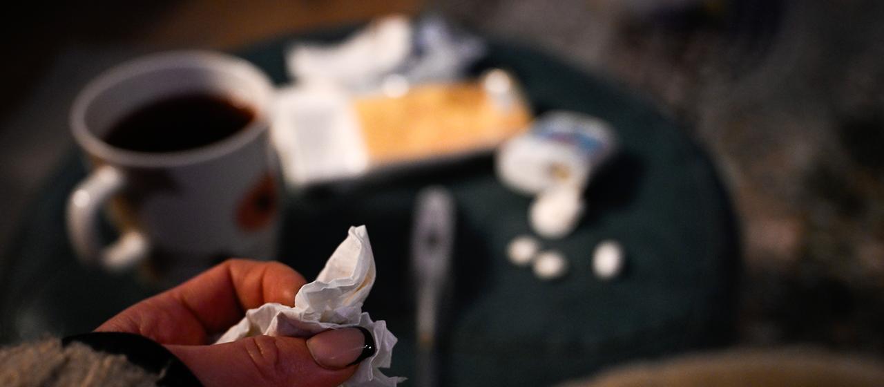 Eine Hand mit einem Taschentuch ist vor einem Tisch mit Tee, Fieberthermometer und Medikamenten zu sehen.