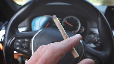 Eine Hand mit Joint hält das Lenkrad eines Autos.
