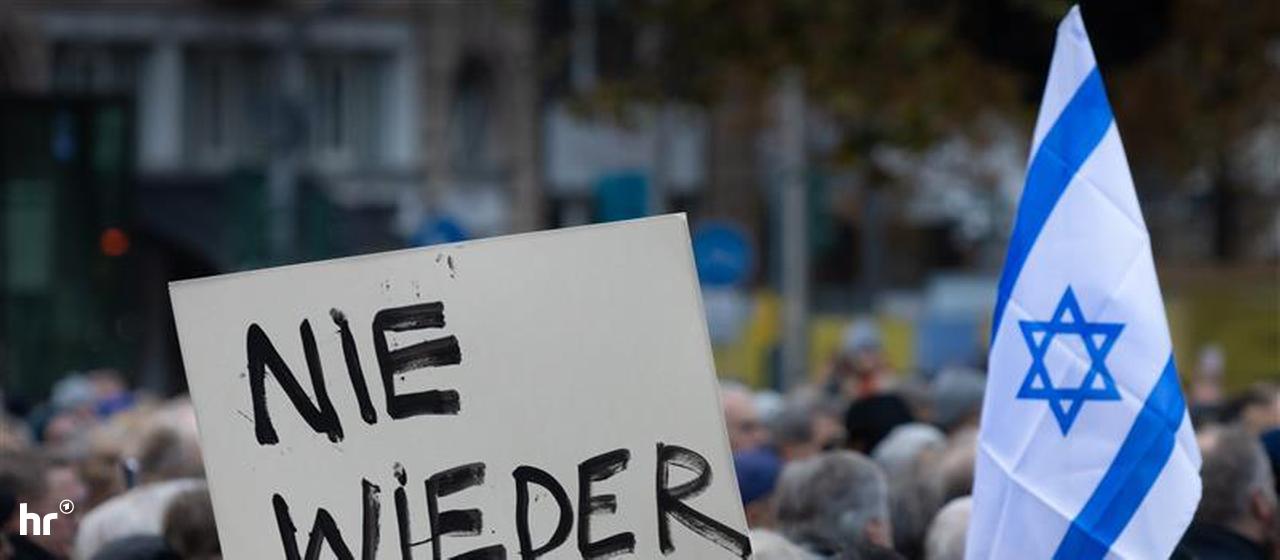 Demonstranten-Köpfe von hinten, es wird eine Israel-Flagge hochgehalten und ein Schild mit der Aufschrift "Nie Wieder"