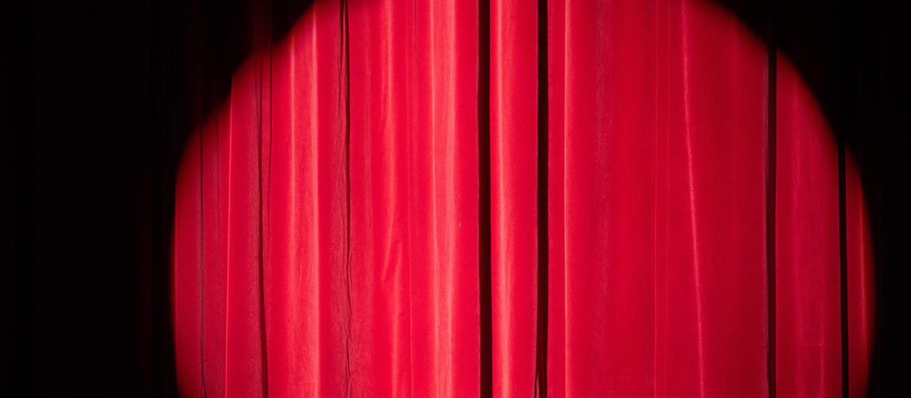 Innenaufnahme einer Zirkusarena. Starke Scheinwerfer und Nebel lassen im Hintergrund Umrisse mehrerer Personen vor einer beleuchteten Leinwand erkennen.