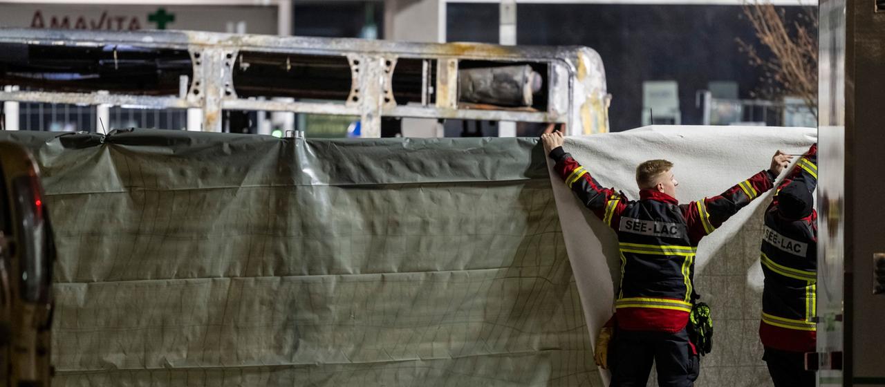Feuerwehrleute verdecken den ausgebrannten Bus mit einer Plane