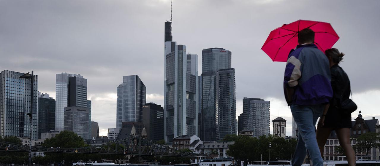 Zwei Personen spazieren vor der Frankfurter Bankenskyline am Mainufer entlang.