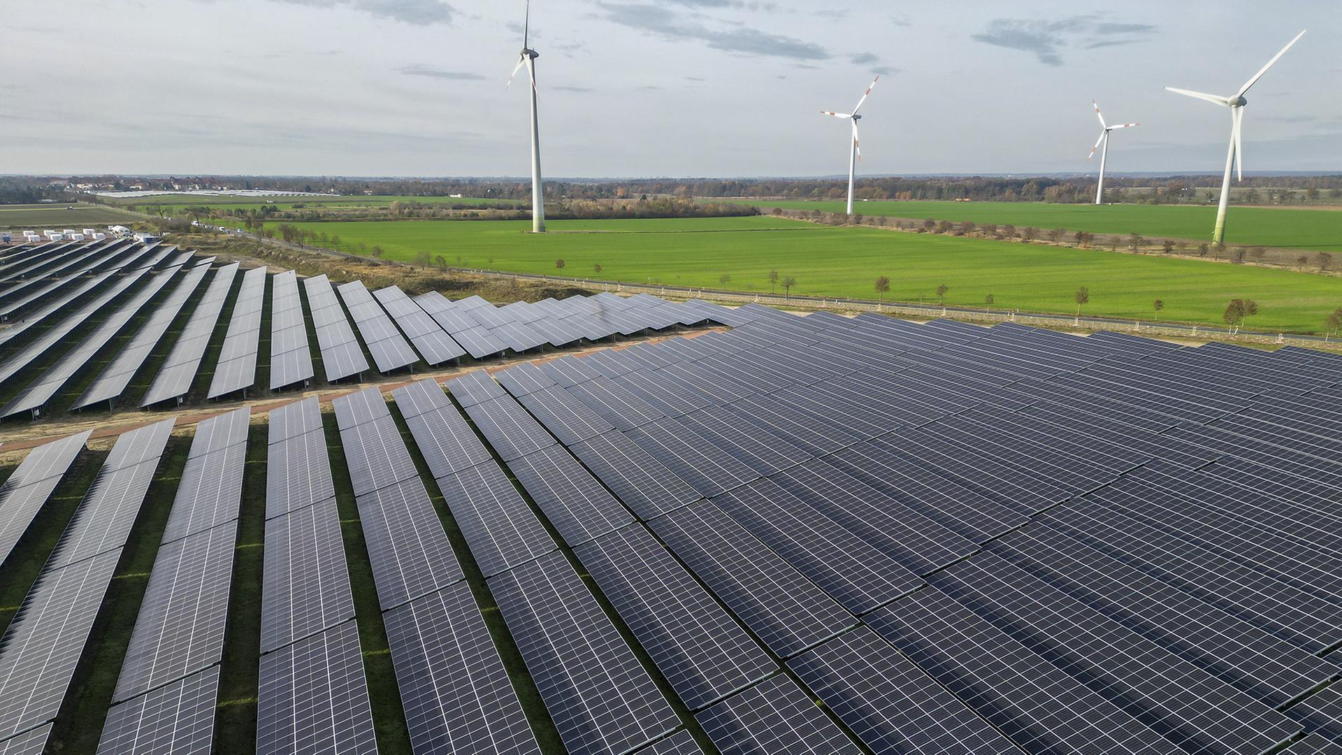 Windkraftanlagen drehen sich hinter den Solaranlagen des PV-Batteriespeicher-Hybridkraftwerks in Zerbst. | dpa