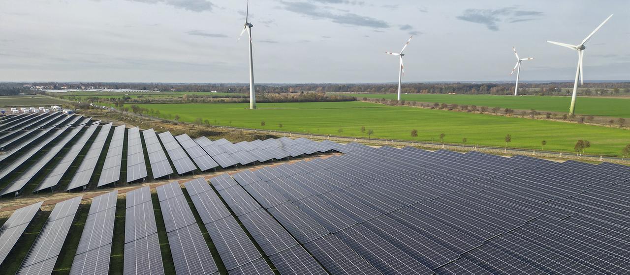 Windkraftanlagen drehen sich hinter den Solaranlagen des PV-Batteriespeicher-Hybridkraftwerks in Zerbst.