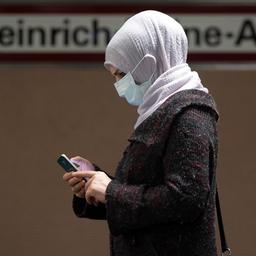 Eine Frau steht mit Mundschutz am Bahnsteig und bedient ihr Smartphone