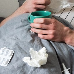 Eine Person liegt mit Teetasse, Taschentüchern und Medikamenten im Bett.